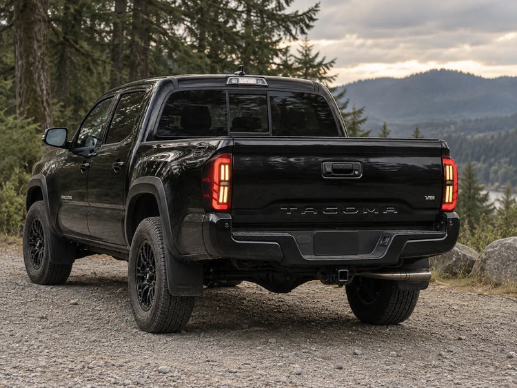 Black Toyota Tacoma parked on a forest road with red and amber LED tail lights illuminated.