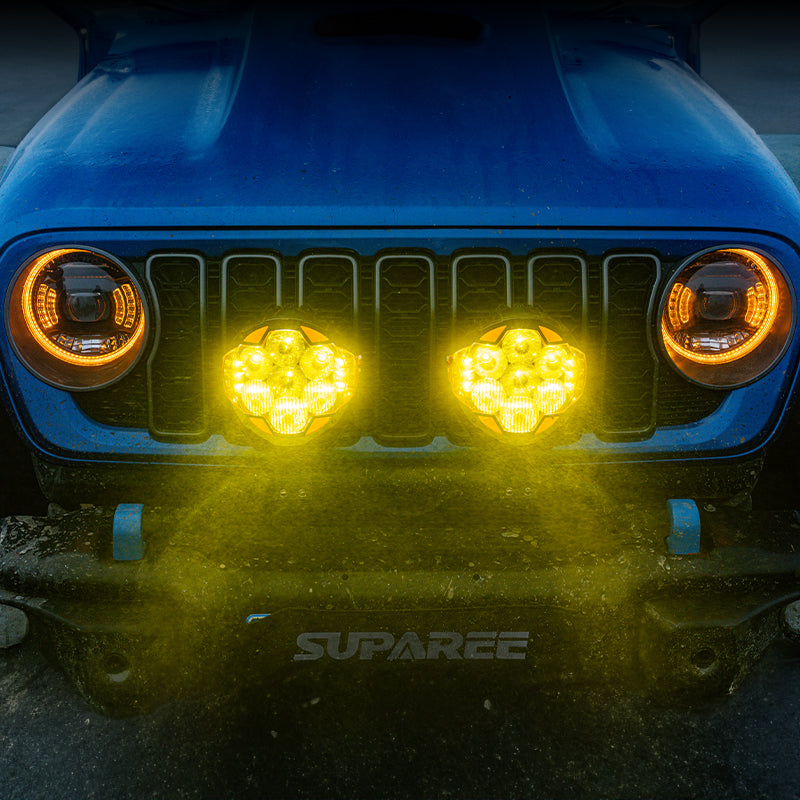 Two SUPAREE HELM 7 LED pod lights shining amber light from the front bumper of a blue Jeep.