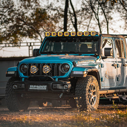 SUPAREE HELM 7 off-road lights mounted below the grille of a blue Jeep parked beside trees.