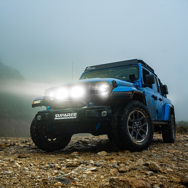 White beams from SUPAREE HELM 7 LED driving lights on a blue Jeep parked on rocky ground in mist.