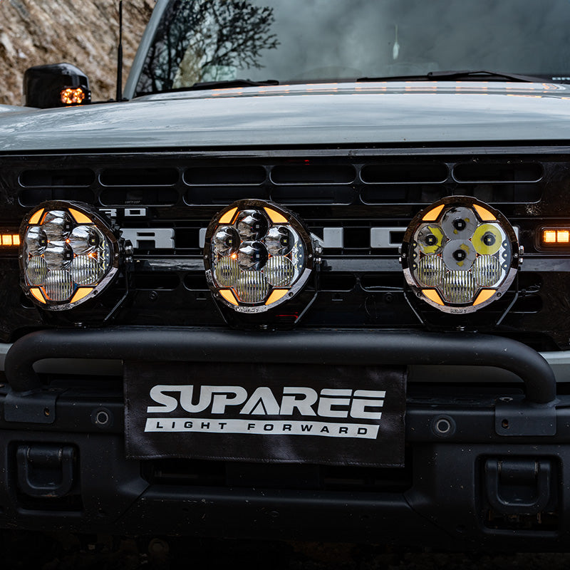 Three round SUPAREE HELM 7 off-road lights mounted across the front bumper of a Ford Bronco.