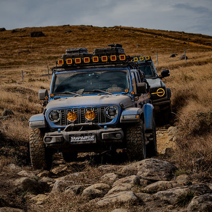 Jeep leading an off-road convoy on a rocky grassland trail with two amber-covered SUPAREE HELM 7 lights on its bumper.