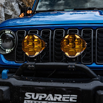 Two SUPAREE HELM 7 LED pod lights with amber covers mounted below the grille of a blue Jeep.