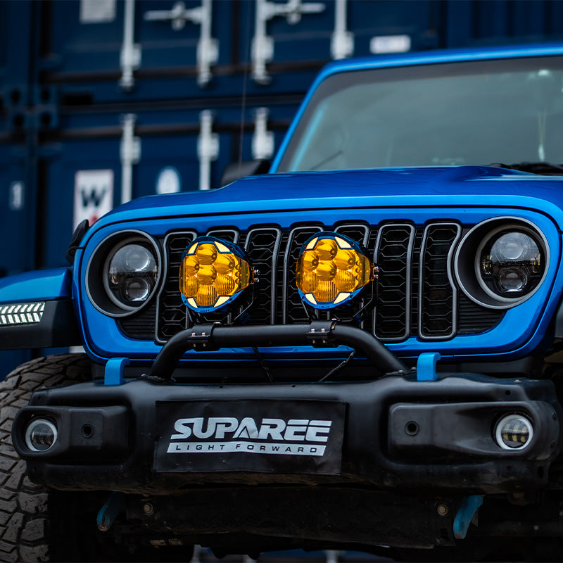Blue Jeep parked beside shipping containers with two amber-covered SUPAREE HELM 7 LED pod lights on its bumper.